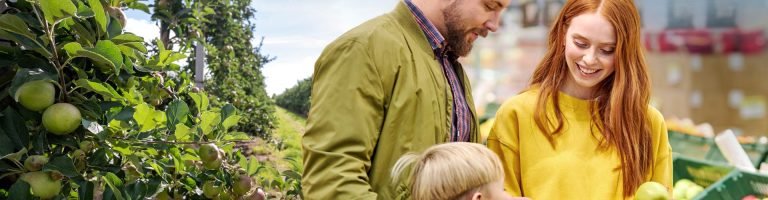 Eine Familie steht an einem Obstregal und vergleicht Äpfel.