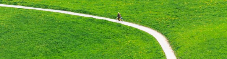 Eine Person fährt mit einem Fahrrad einen langen Weg zwischen zwei großen Wiesen entlang.